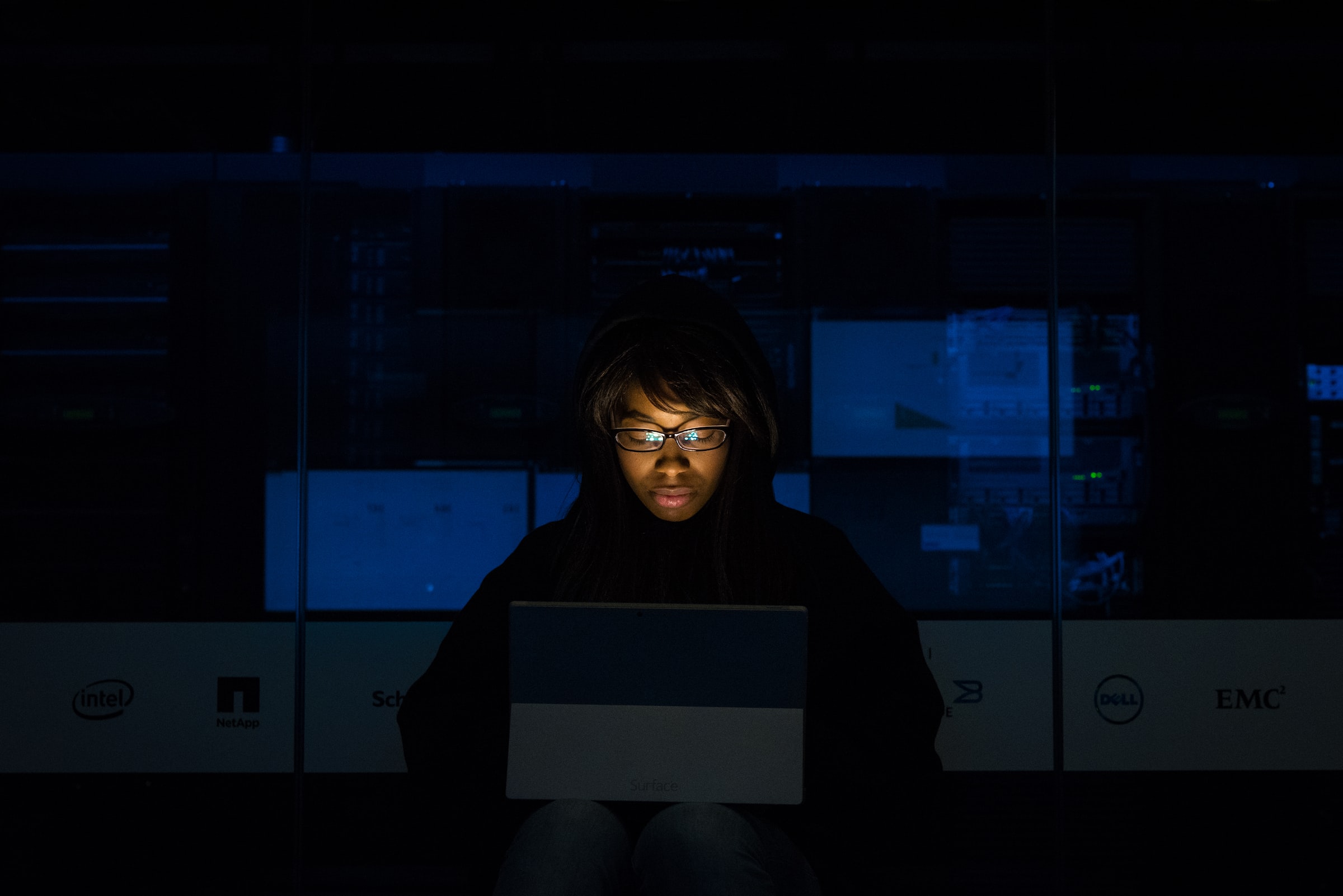 Vemos uma mulher negra concentrada enquanto trabalha em seu computador numa sala escura (imagem ilustrativa).
